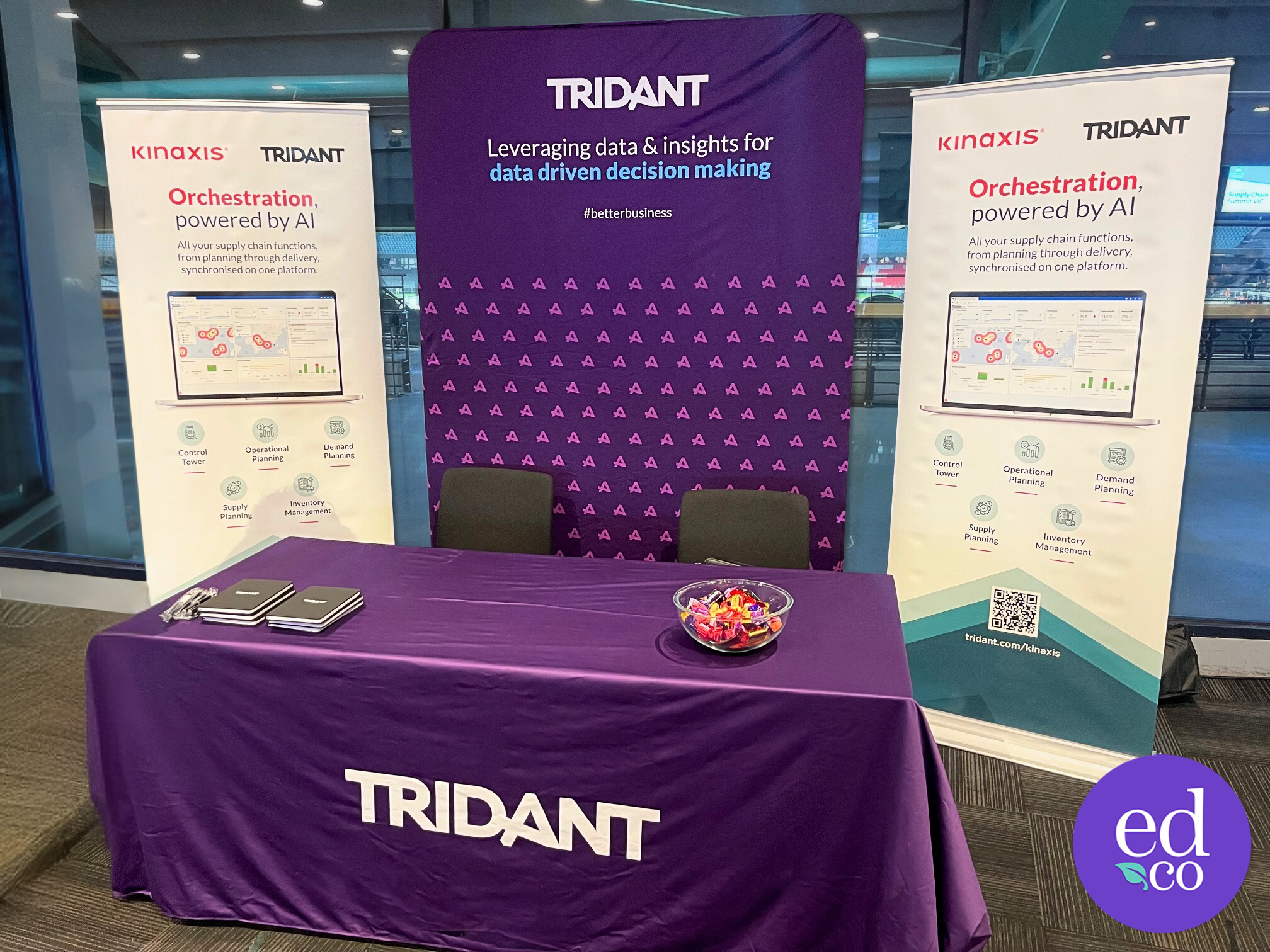 Photograph of Tridant exhibition stand with purple banners designed by EDCo., promoting Kinaxis technology for supply chain management and decision-making. EDCo. logo in the bottom left.