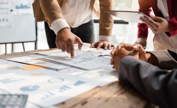 Business team reviewing printed climate-risk charts on a tabletop while discussing data on a tablet — representing collaborative TCFD reporting.